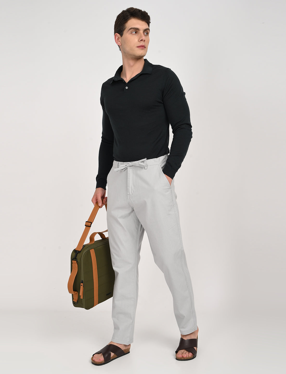 Model wearing light grey linen blend trousers with black long-sleeve polo and holding a green bag.